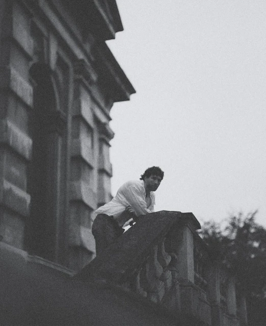 Monochromatic portrait of a man in a white shirt leaning on a stone balcony with trees and an old building in the background.