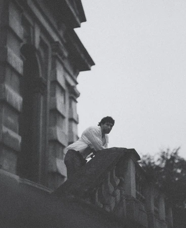 Monochromatic portrait of a man in a white shirt leaning on a stone balcony with trees and an old building in the background.