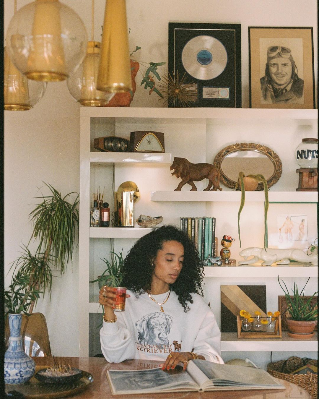 Woman with curly hair sits at wooden table, drinking from a glass, with a photo album and bookshelf visible in the background.