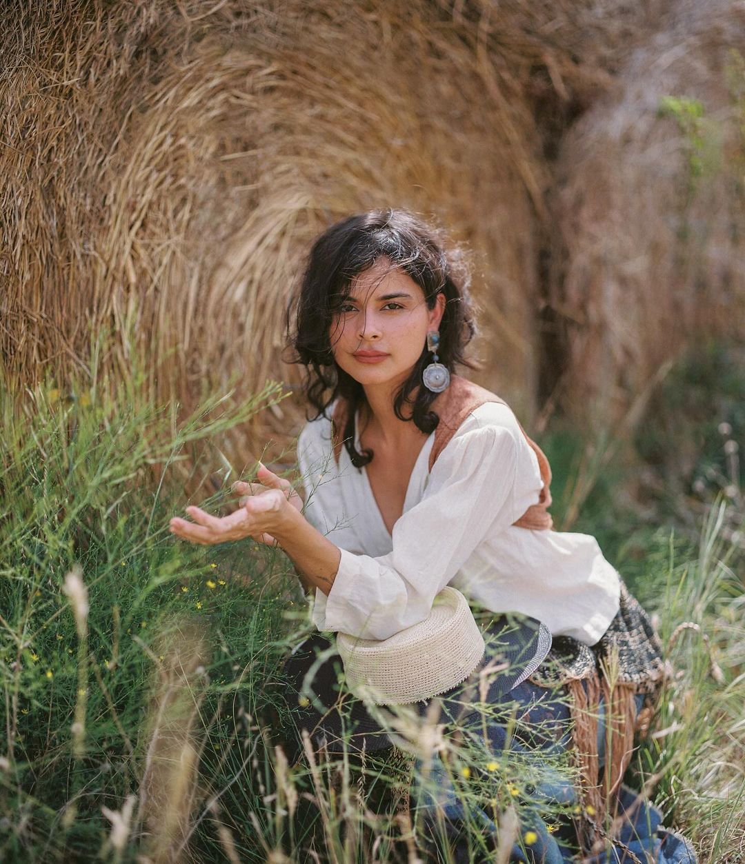 Woman in white blouse poses outdoors near hay bale, wearing large earrings, with green plants in foreground on a sunny day.