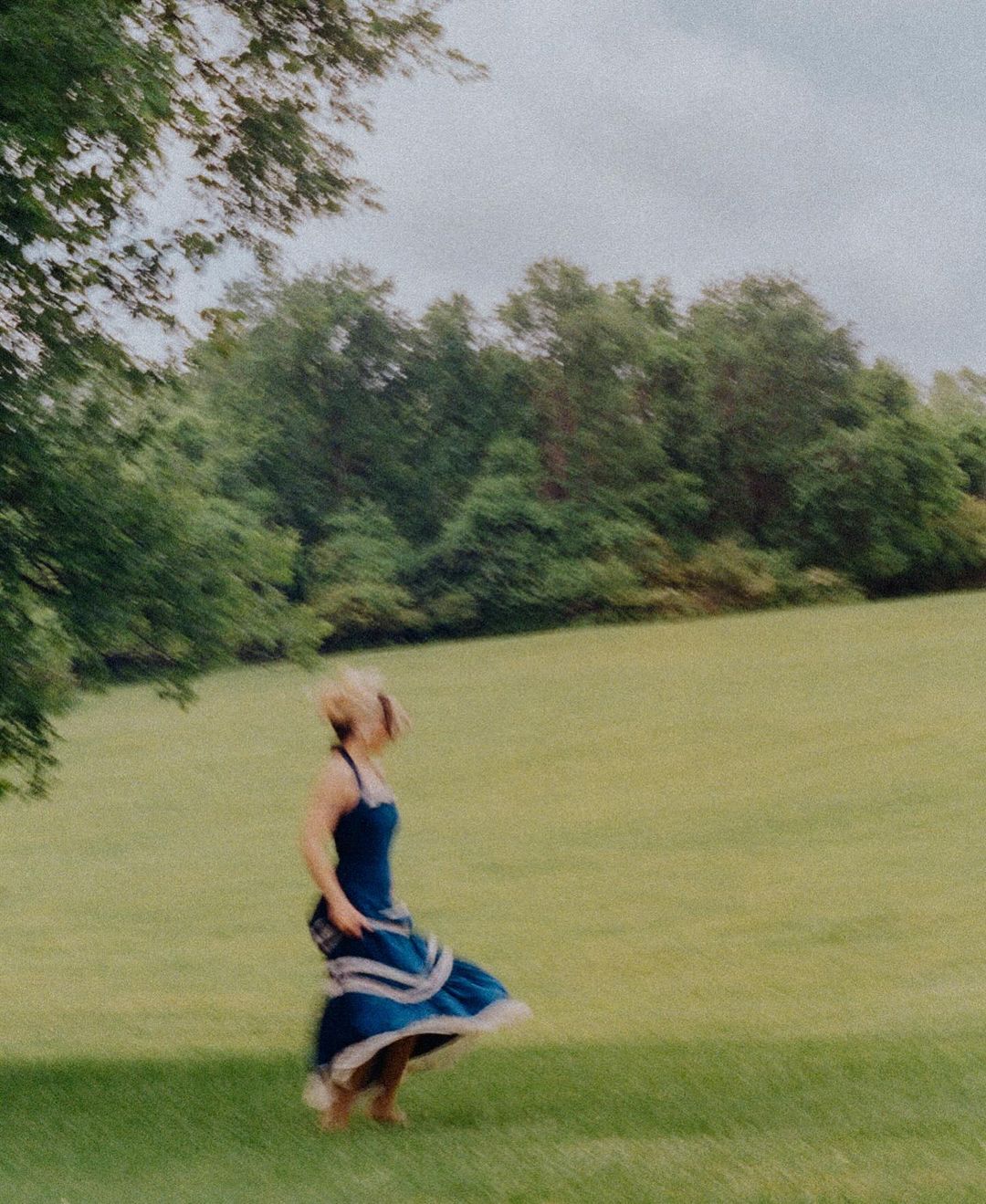 Woman in a blue dress twirls in a grassy field with trees in the background, capturing a sense of movement and freedom in nature.
