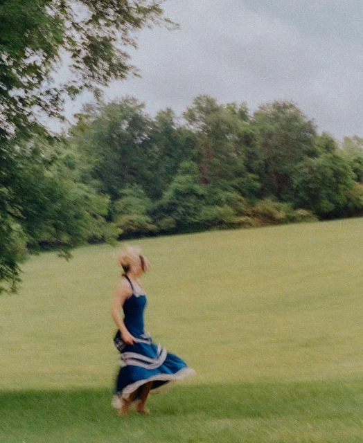 Woman in a blue dress twirls in a grassy field with trees in the background, capturing a sense of movement and freedom in nature.