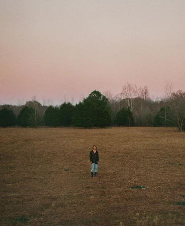 A lone person stands in a vast, golden field under a dusky sky, surrounded by distant trees, creating a serene and expansive scene.