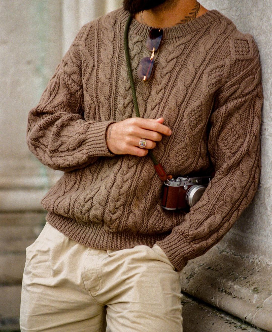 Man wearing a brown cable knit sweater and beige trousers, leaning against a stone wall with a vintage camera.