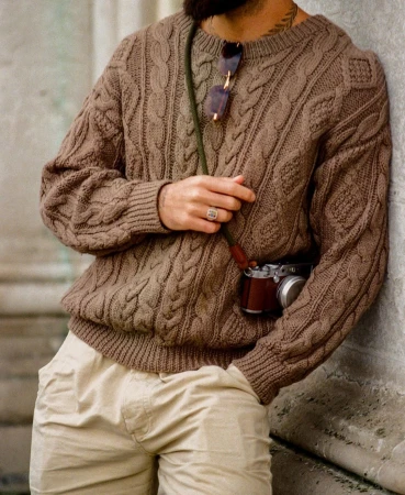 Man wearing a brown cable knit sweater and beige trousers, leaning against a stone wall with a vintage camera.