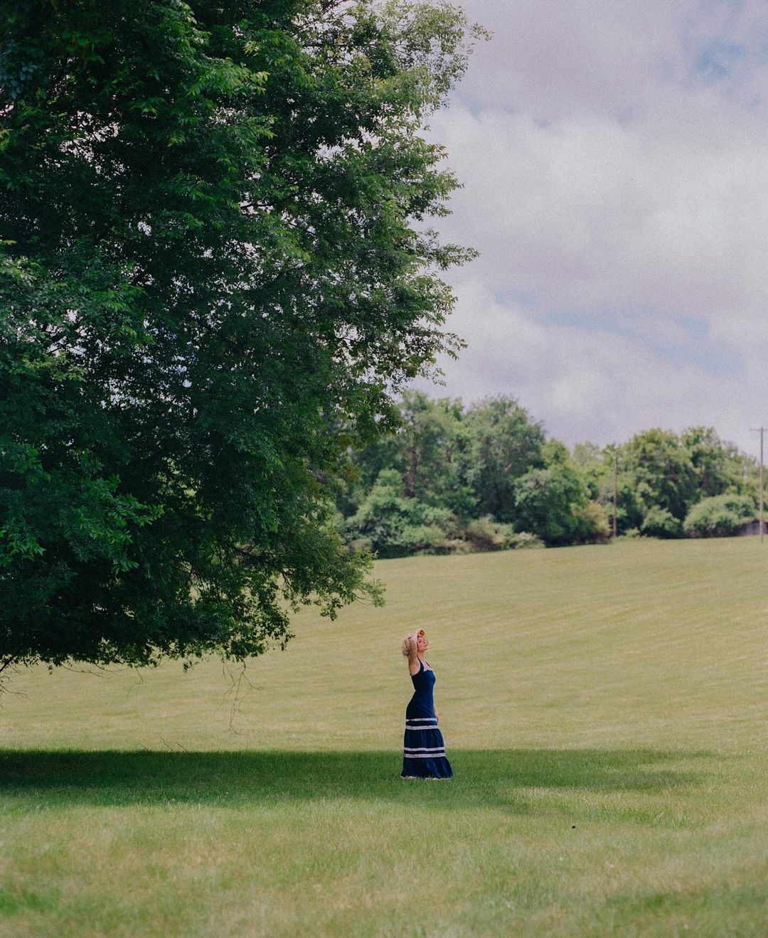 A woman in a blue dress stands beneath a large tree in a grassy field under an overcast sky, captured by Bethany Reed.