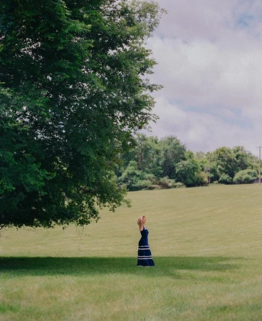A woman in a blue dress stands beneath a large tree in a grassy field under an overcast sky, captured by Bethany Reed.