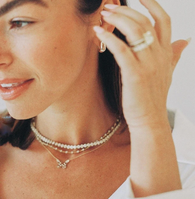 Close up of a woman with long hair, gold earrings, layered pearl necklaces and a butterfly pendant, wearing a white collared shirt.