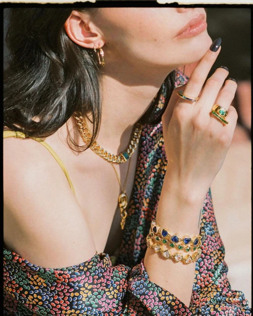 Woman displays luxury gold jewelry including bracelets, rings, necklace and earrings in a studio setting with diffused lighting.