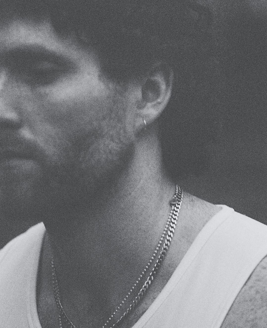 Monochromatic close-up of a light-skinned adult male with a white tank top and silver chain necklaces. Moody lighting, shallow depth of field.