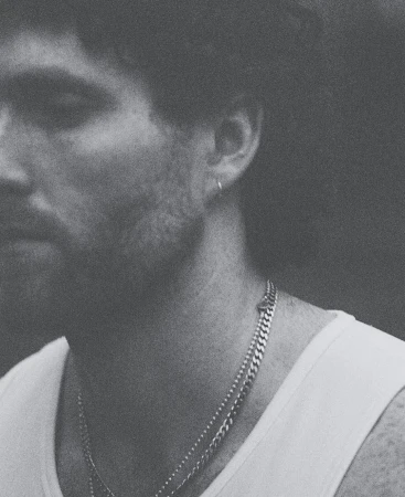 Monochromatic close-up of a light-skinned adult male with a white tank top and silver chain necklaces. Moody lighting, shallow depth of field.