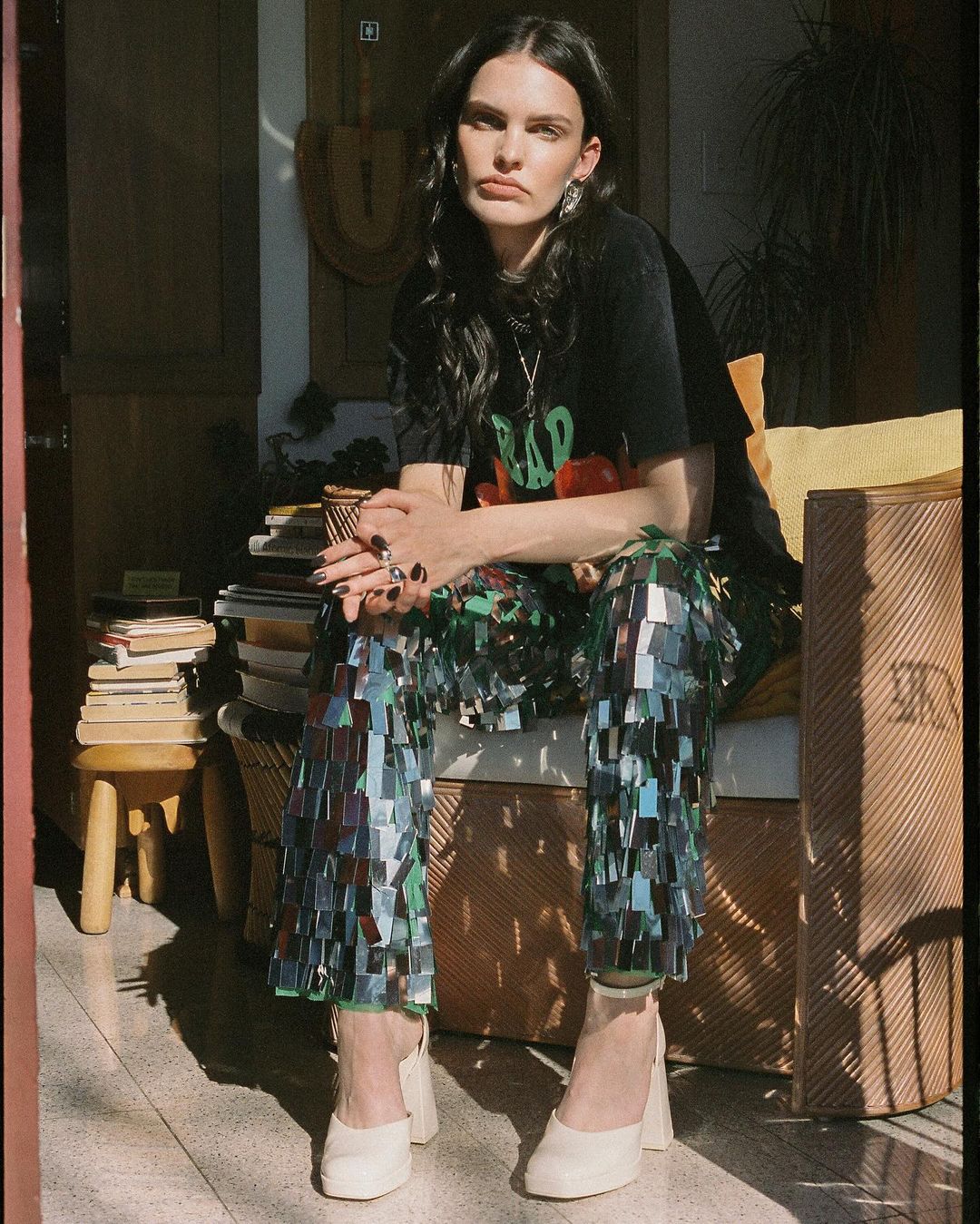 Indoor portrait of a woman in a t-shirt and shiny pants, seated on a wicker chair near a stack of books, with natural sunlight.