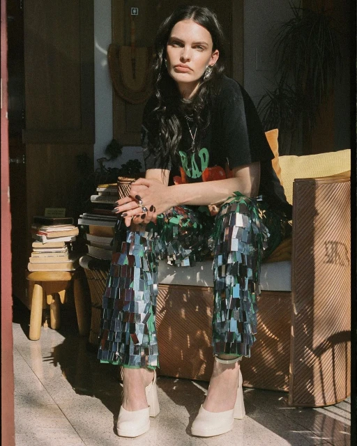 Indoor portrait of a woman in a t-shirt and shiny pants, seated on a wicker chair near a stack of books, with natural sunlight.