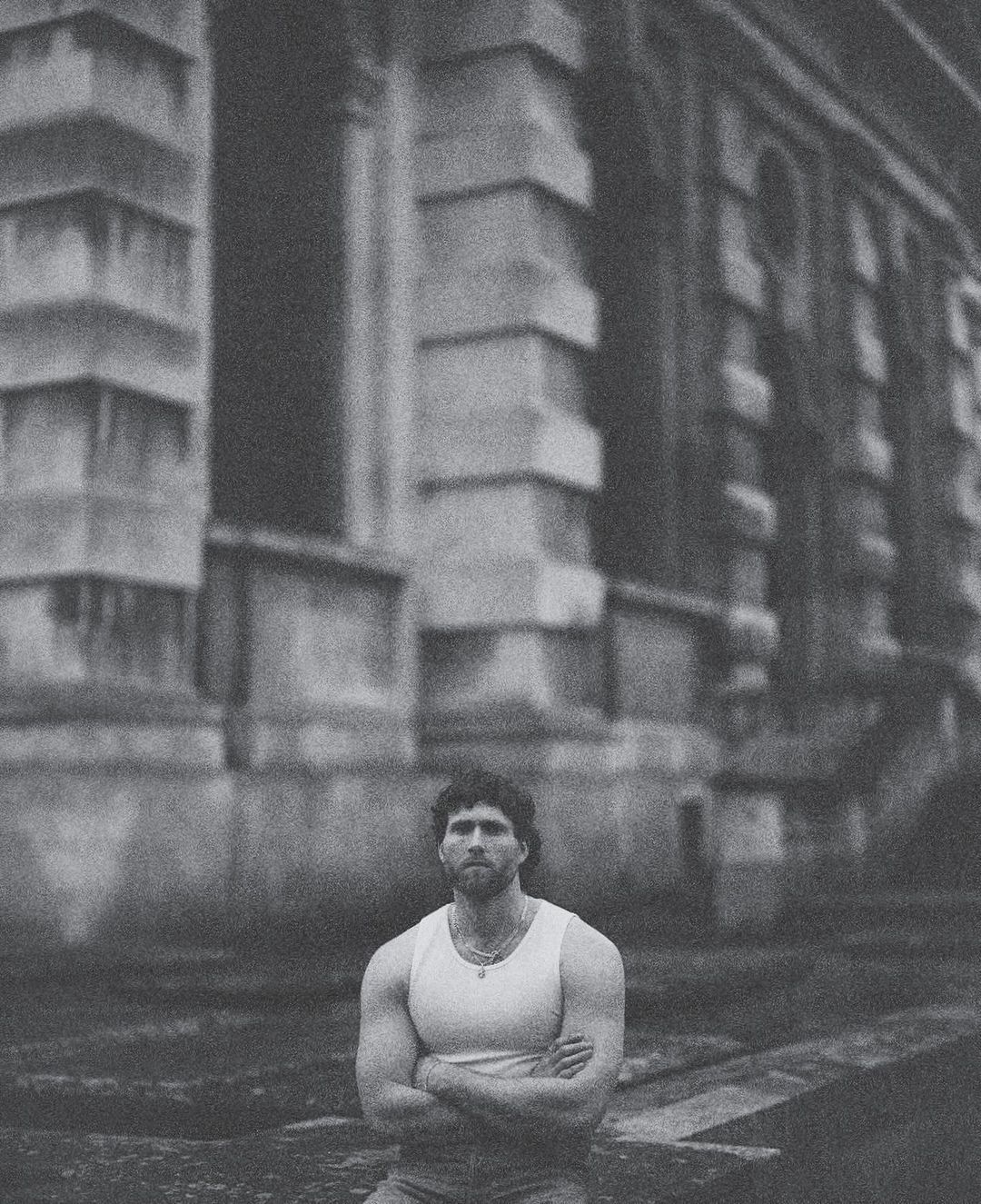 Man with arms crossed wearing a white tank top outdoors against classical architectural building exterior