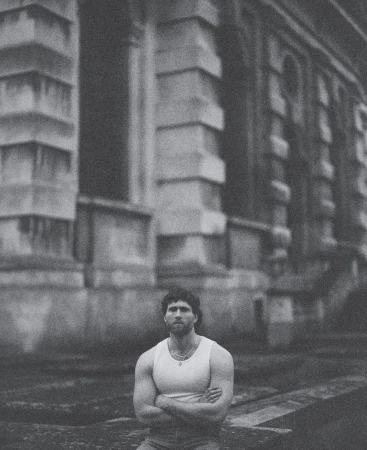 Man with arms crossed wearing a white tank top outdoors against classical architectural building exterior
