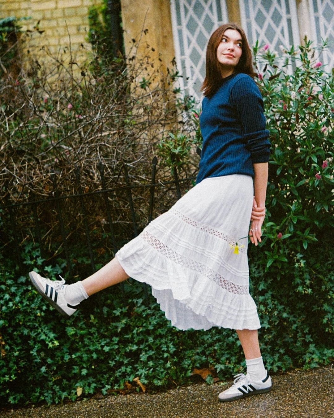 Woman in dark blue sweater and white skirt lifting her leg outdoors by green bushes