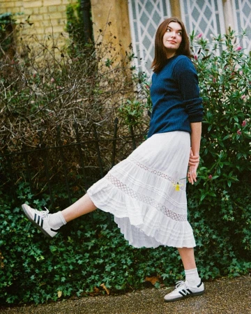 Woman in dark blue sweater and white skirt lifting her leg outdoors by green bushes