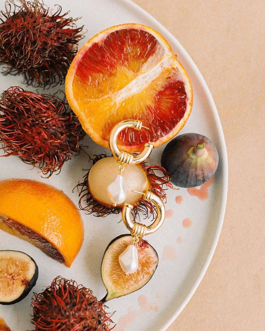 Still life of a plate of sliced blood oranges, rambutans, and figs, adorned with gold hoop earrings featuring baroque pearl pendants.