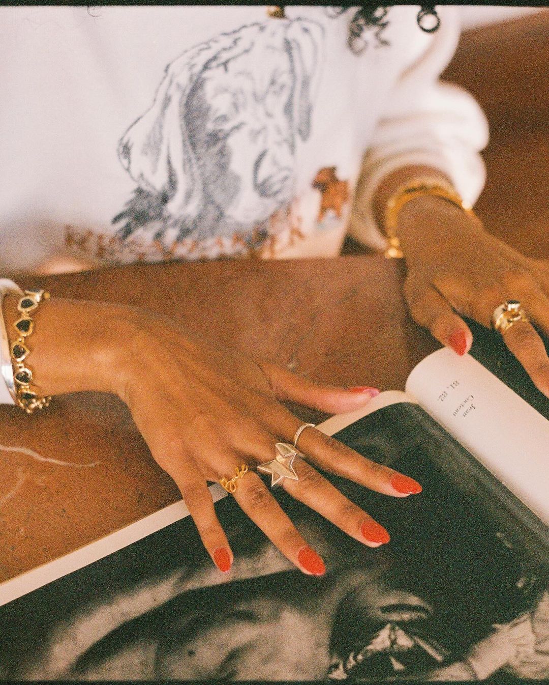 Person's hands with red nail polish and gold rings are resting on an open book. Close-up studio shot with fine art aesthetic.