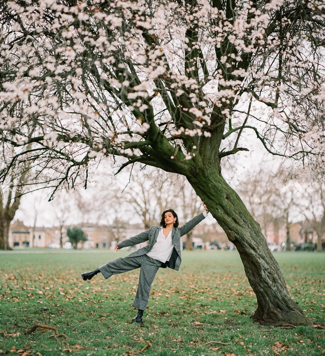 Young woman in gray suit balances near a blooming tree in a park, showcasing a playful pose and urban outdoor setting.