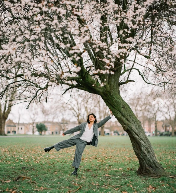 Young woman in gray suit balances near a blooming tree in a park, showcasing a playful pose and urban outdoor setting.