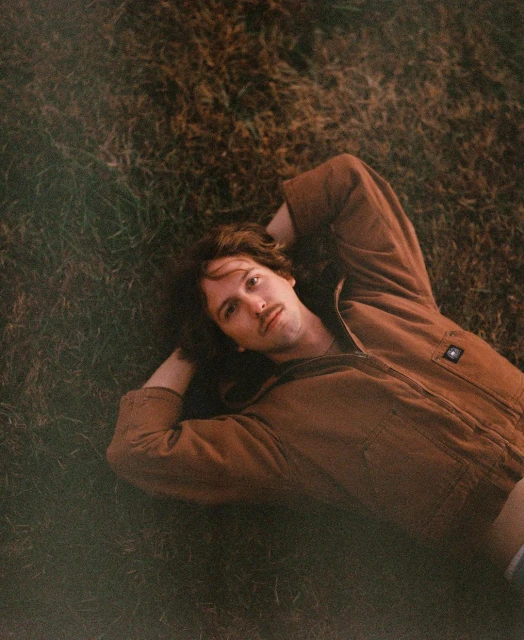 A male with brown hair and a mustache lying on a grassy surface, looking at the camera with hands behind his head.