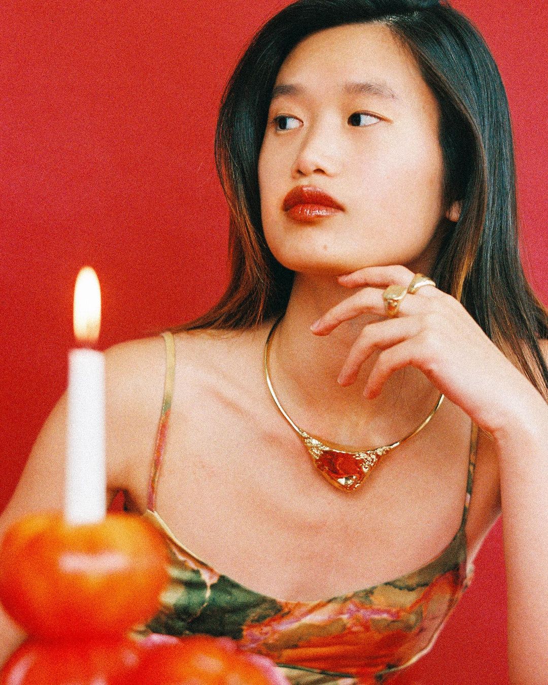 Portrait of an Asian woman with dark hair and red lipstick wearing a gold necklace with a red gem, with a candle and red background.