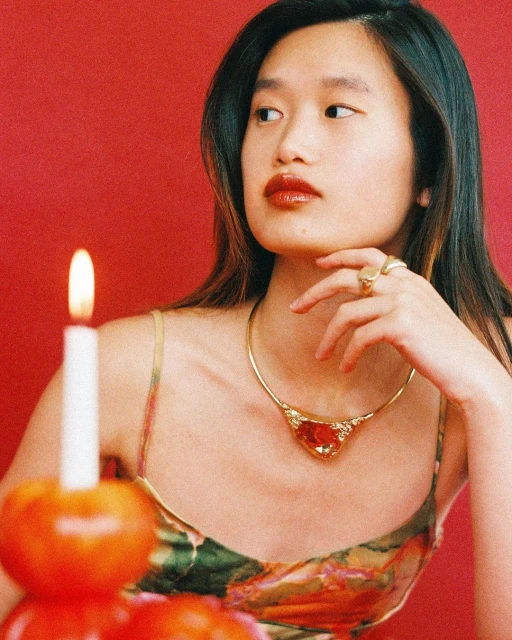 Portrait of an Asian woman with dark hair and red lipstick wearing a gold necklace with a red gem, with a candle and red background.
