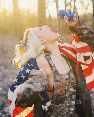 Woman in camouflage overalls and draped American flag drinking from a blue can outdoors in sunlight.