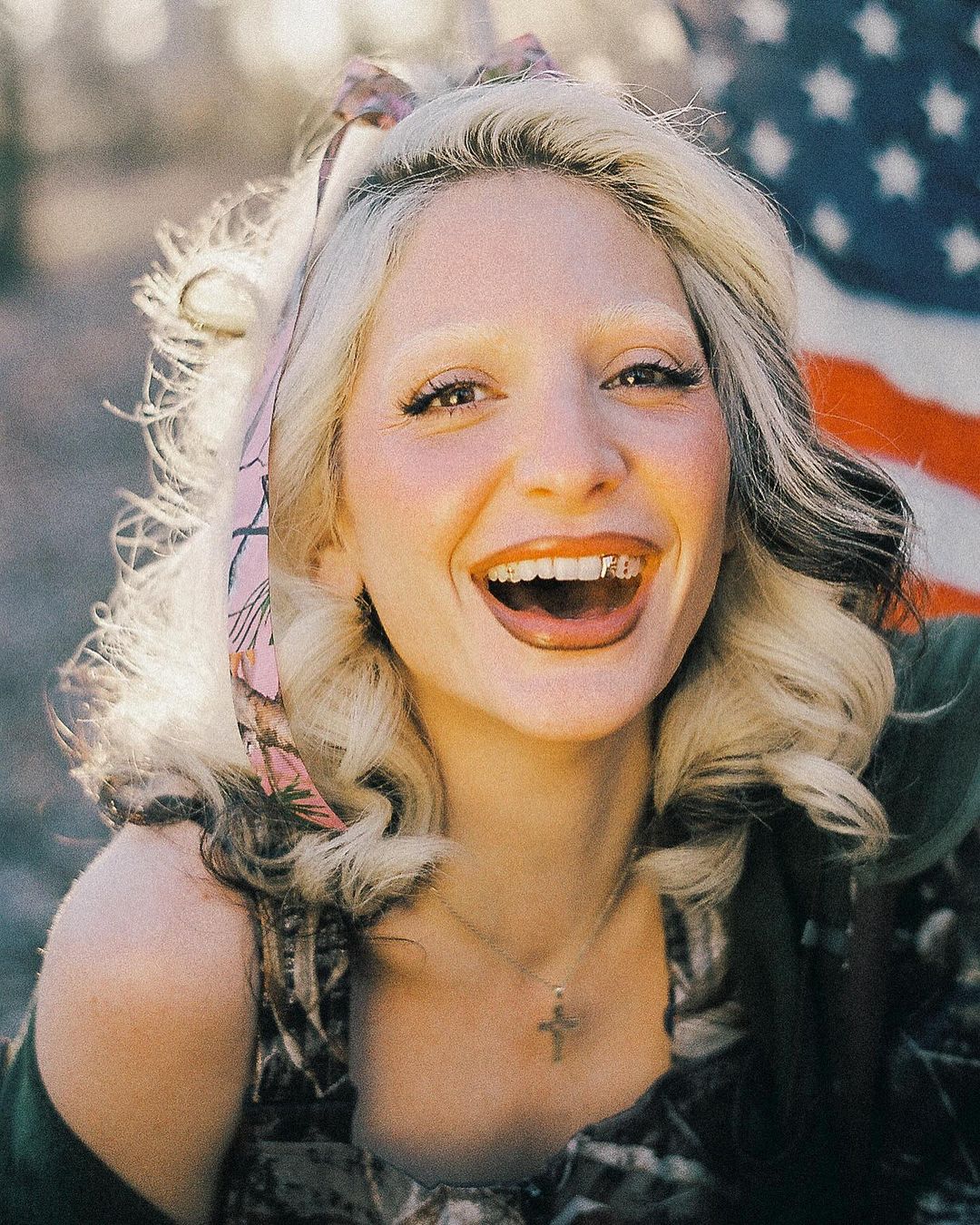 Smiling blonde woman with a floral headband in a close-up portrait, with an American flag blurred in the background.