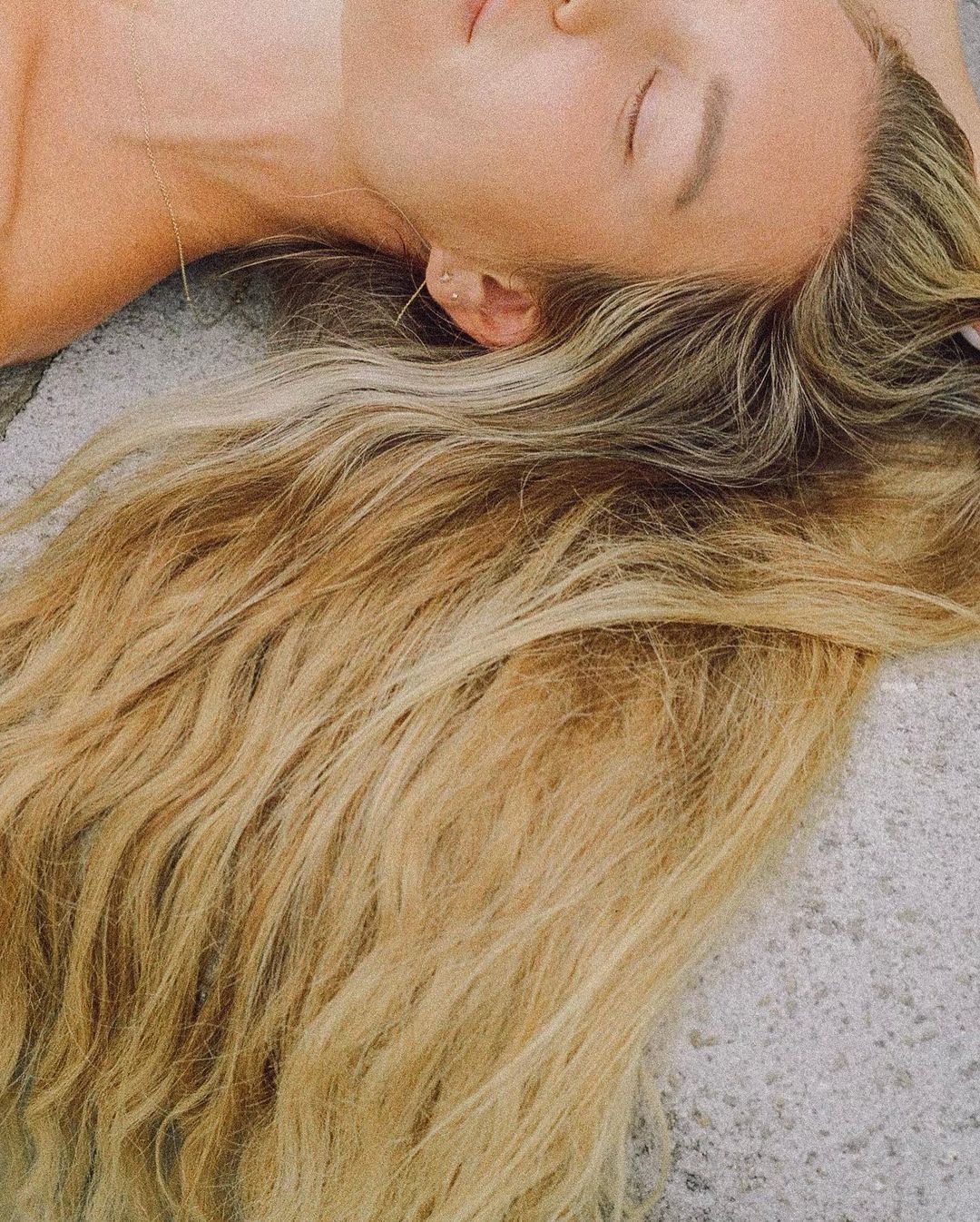 Close-up of a fair-skinned adult female laying down with her eyes closed and long, blond wavy hair fanned out over a gray surface.