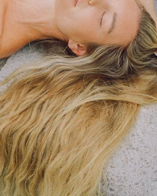 Close-up of a fair-skinned adult female laying down with her eyes closed and long, blond wavy hair fanned out over a gray surface.