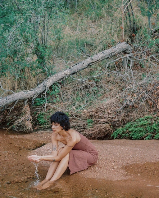 Woman in dress sits at edge of water in an outdoor nature setting with a fallen tree. She holds water in her hands.