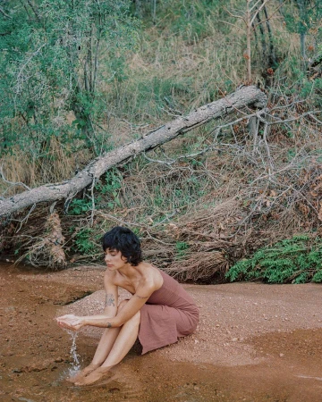 Woman in dress sits at edge of water in an outdoor nature setting with a fallen tree. She holds water in her hands.