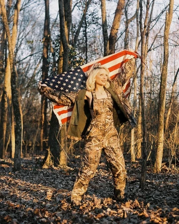 Woman in camo overalls stands in a forest holding an American flag draped over her shoulders, backlit by natural light.