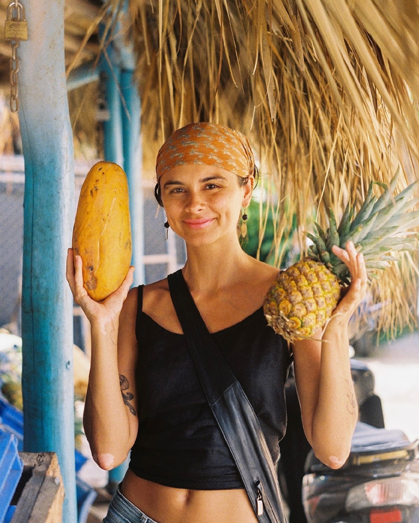 Smiling woman holds a papaya and pineapple in front of a thatched-roof structure, wearing an orange headscarf and black top.