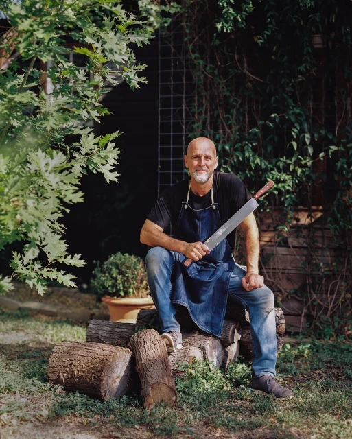 Bald adult male sitting on logs in an outdoor setting, wearing an apron and holding a large blade. Natural light and foliage.