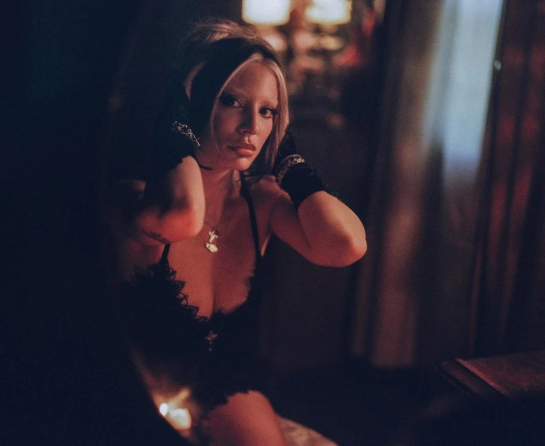 Blonde woman in a dark room, wearing a black top, a necklace, and wristbands, looking at the camera with a neutral expression.