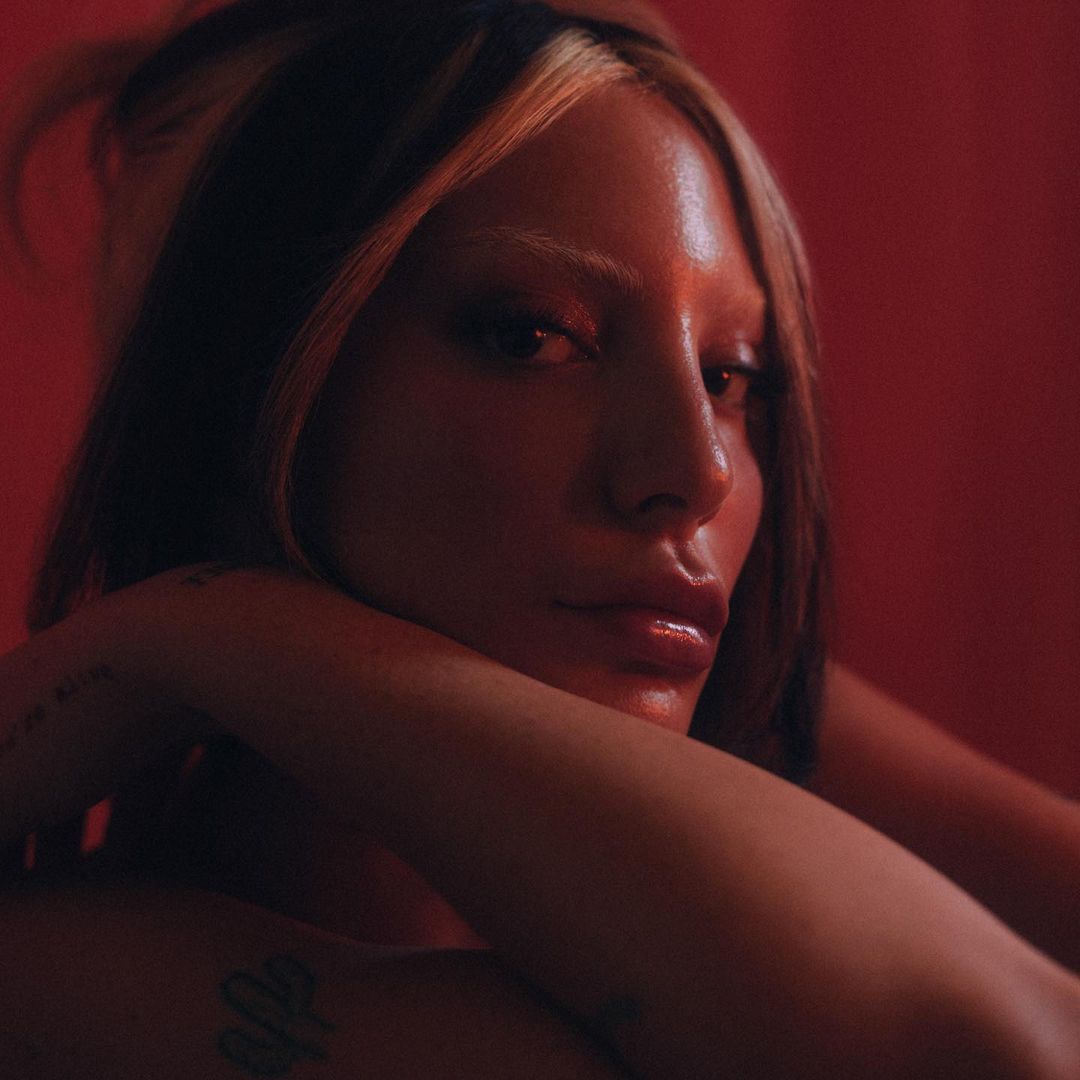 Portrait of a woman with blonde-streaked hair and glossy lips, arms crossed, against a solid red backdrop, illuminated by soft, diffused lighting.