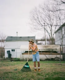 Young man wearing a bucket hat and denim shorts standing in a yard holding a rake by photographer Bethany Reed.