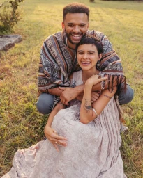 Smiling man and woman outdoors on a grassy lawn, with the man embracing the woman from behind in a patterned poncho.