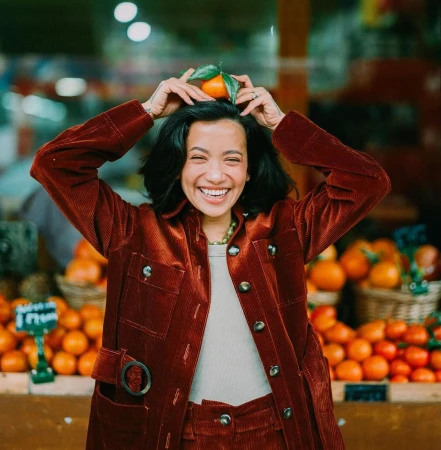 Smiling woman in a rust corduroy jacket balancing a small orange with green leaves on her head at a market