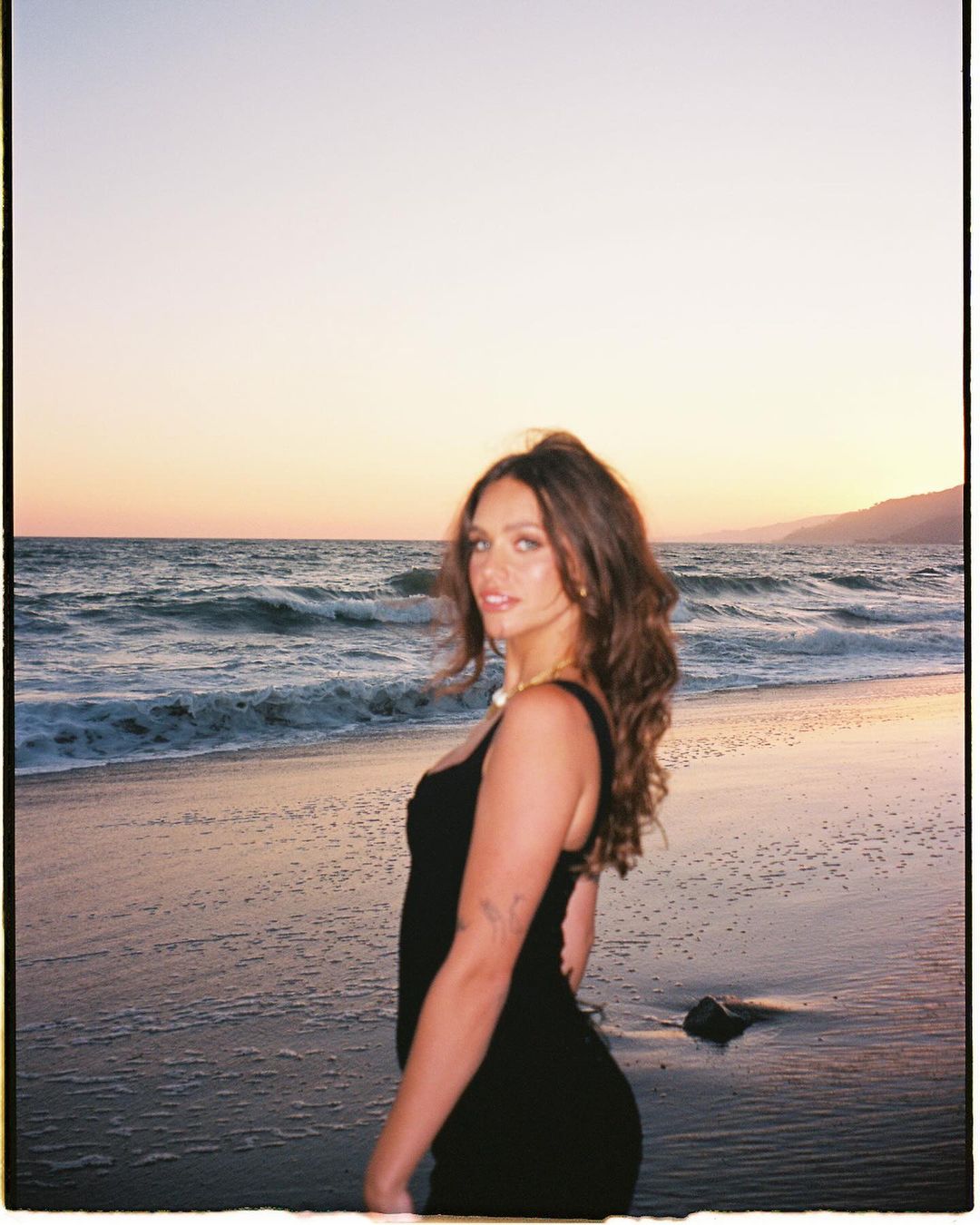 A woman in a black dress standing on a sandy beach at sunset by the ocean, captured by photographer Bethany Reed.