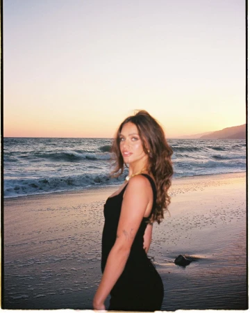 A woman in a black dress standing on a sandy beach at sunset by the ocean, captured by photographer Bethany Reed.