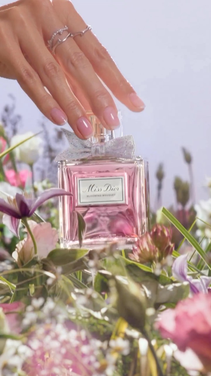 Close-up shot of a Miss Dior perfume bottle surrounded by pink and white flowers, with a manicured hand gently touching the bottle.