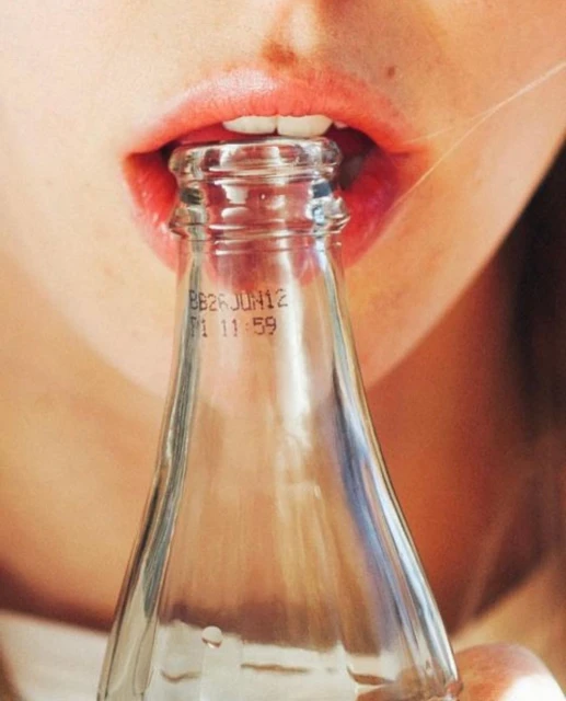 Close-up of a person drinking from a clear glass bottle, highlighting the bottle's opening and the person's lips.