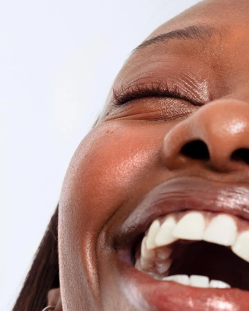 Close-up portrait of a smiling adult woman with dark skin and an open mouth, revealing her teeth, against a clean white background.