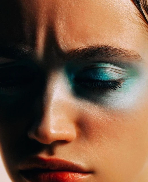 Close-up beauty shot of a woman with closed eyes and bold teal and white eyeshadow, partially obscured by dark shadow.