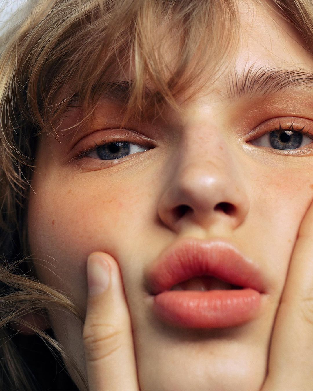 Close-up portrait of a young adult female with blonde hair and blue eyes touching her face in a beauty or fashion shot.