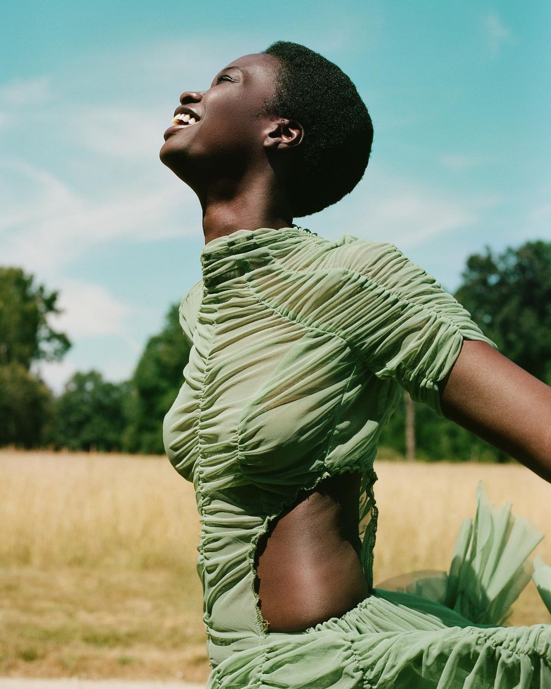 Young adult Black woman in sheer, ruched light-green dress looks upward in sunlit field with trees under blue sky on sunny day.